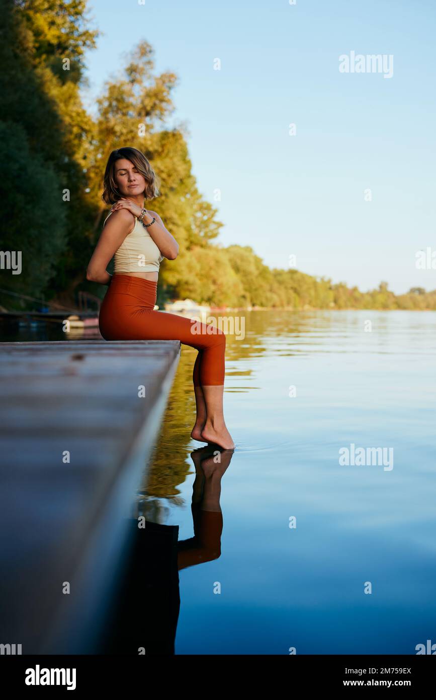 A middle-aged woman is relaxing on the dock and doing yoga breathing exercises. She put her feet in the water. Stock Photo
