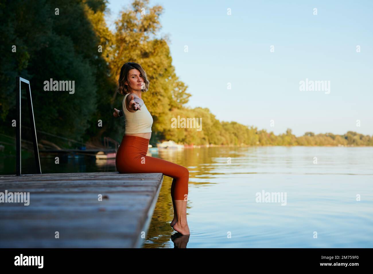 A middle-aged woman is relaxing on the dock and doing yoga breathing exercises. She put her feet in the water. Stock Photo
