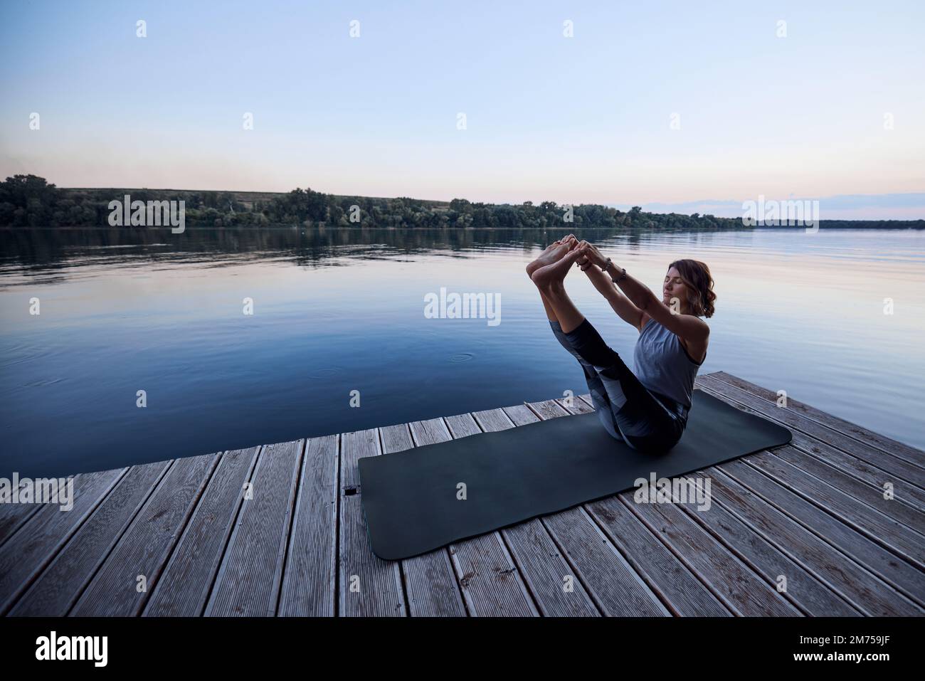 A fit yogi woman is practicing yoga on a dock at dusk. She is in the