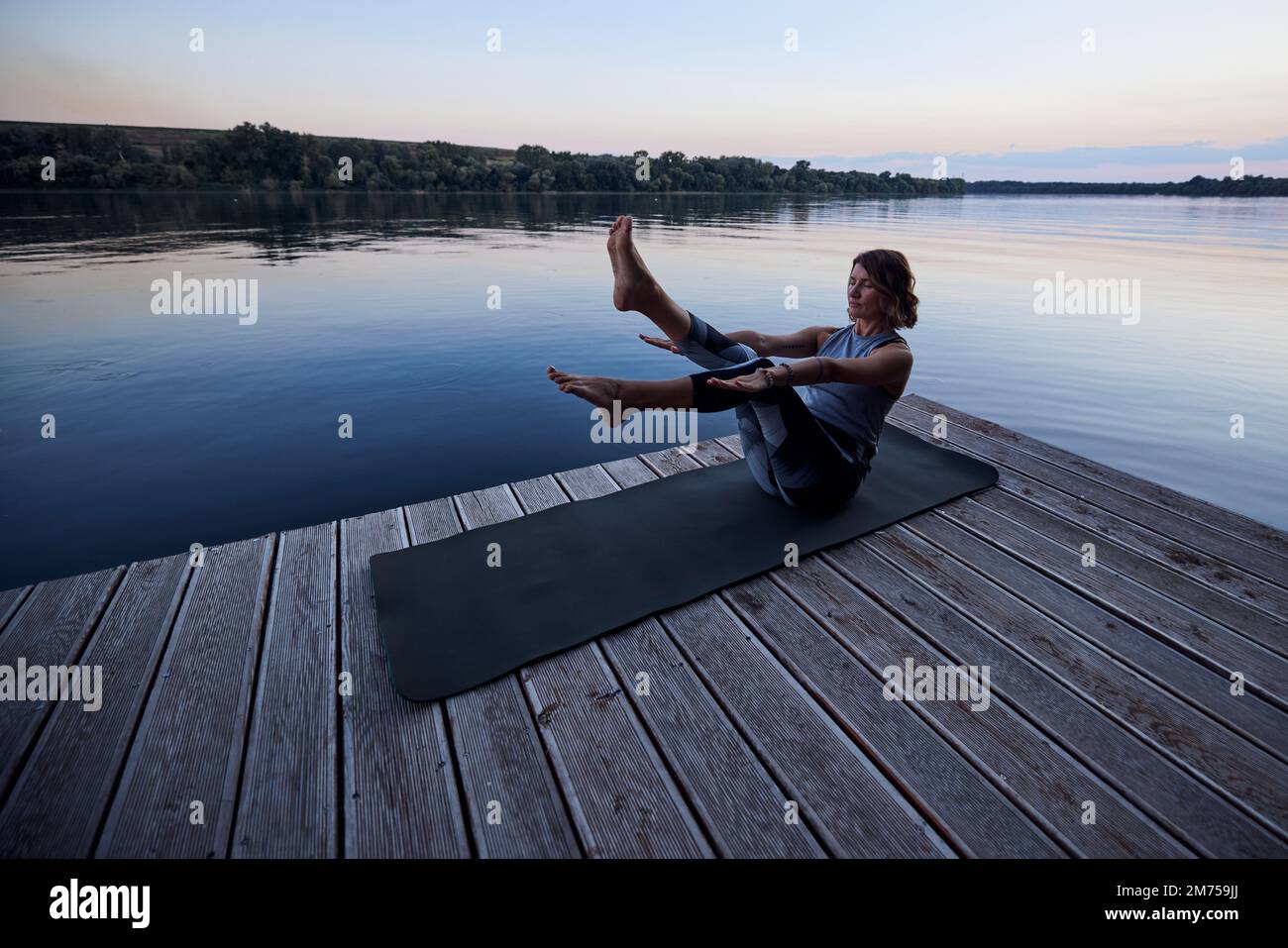 A fit yogi woman is practicing yoga on a dock at dusk. She is in the