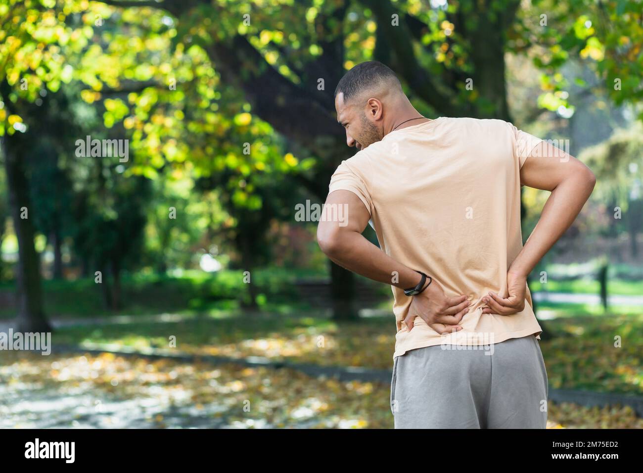 Tired man after jogging in park, hispanic man has severe pain in back and muscles after fitness, massaging his side with hand near trees. Stock Photo