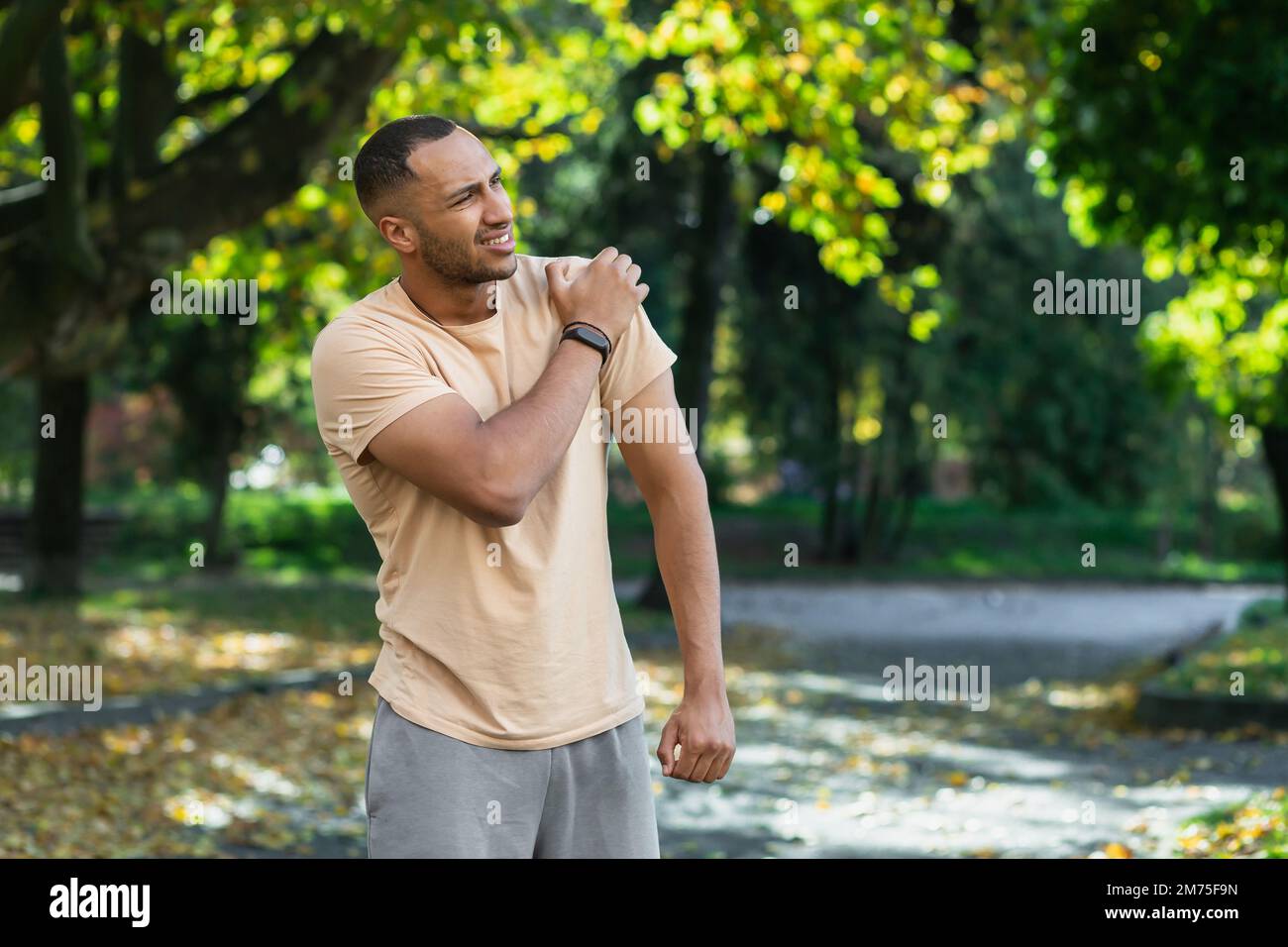 A man injured his shoulder during a fitness class, an African-American man injured himself while jogging in the park, stretches his arm and massages his sore muscles. Stock Photo