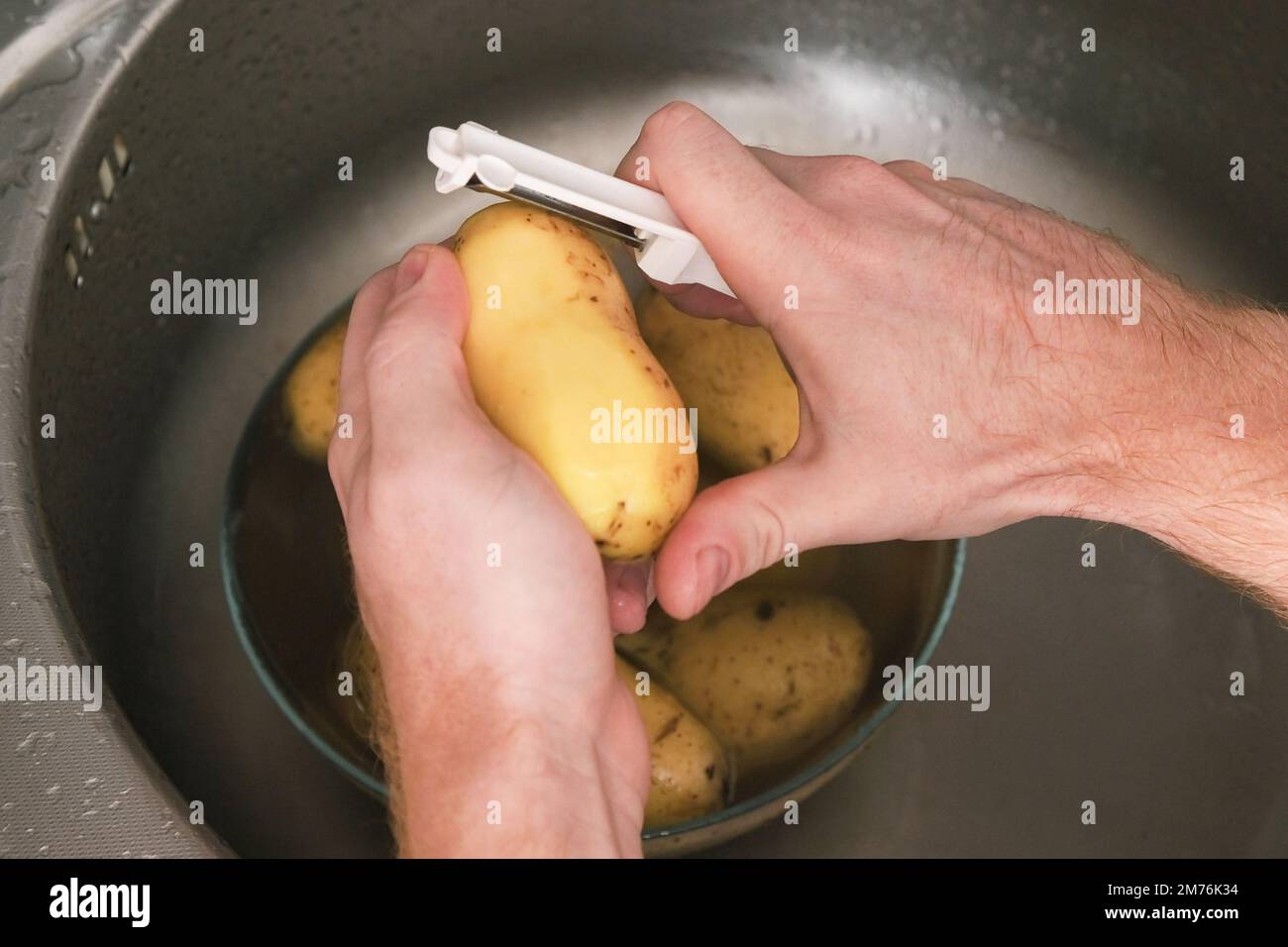 A man peels potatoes with a vegetable peeler in the sink. Preparation