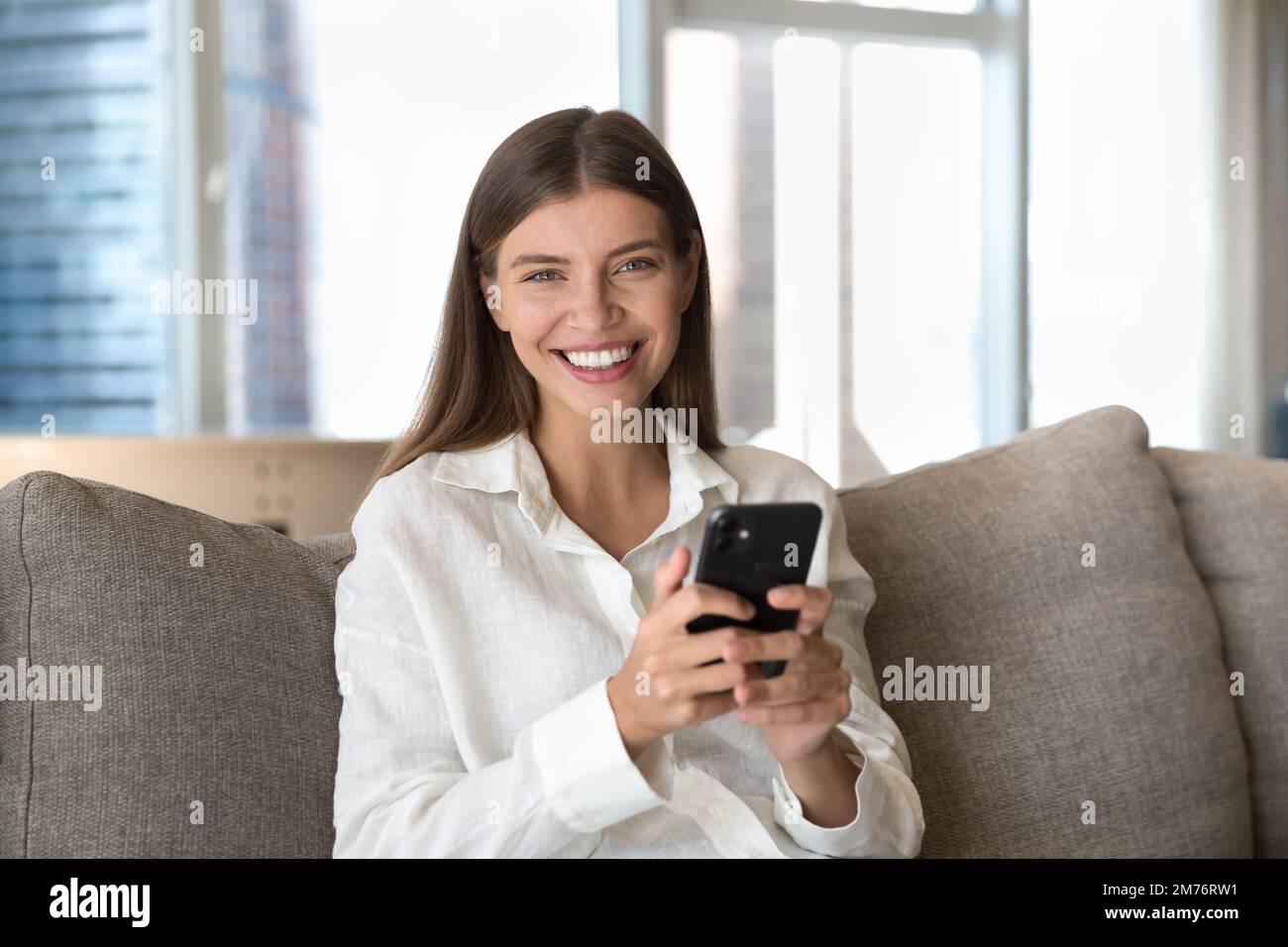 Woman sit on couch with cellphone smile looks at camera Stock Photo
