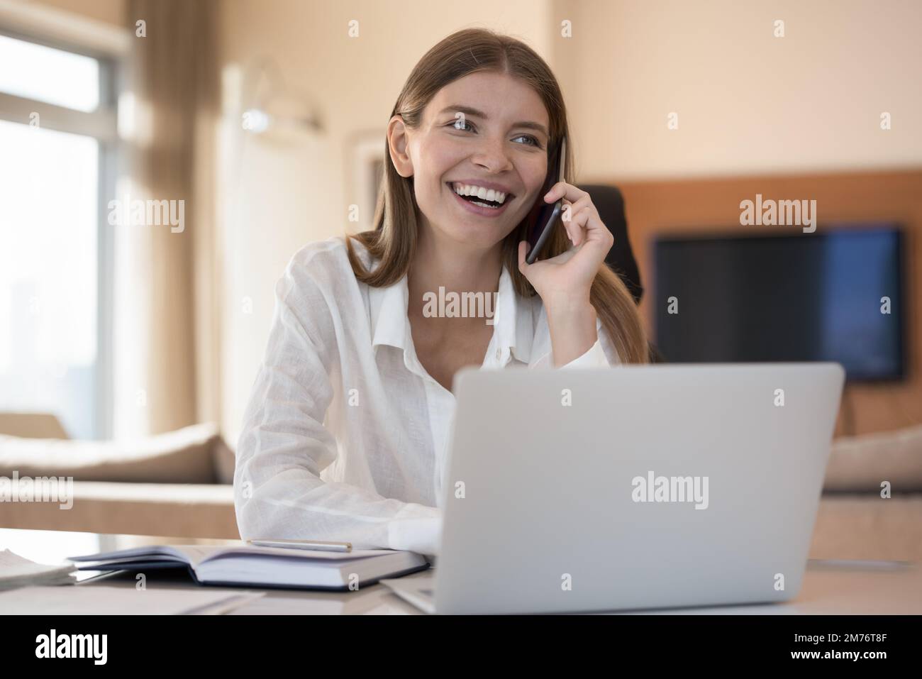 Woman sit at desk with laptop and talks on smartphone Stock Photo