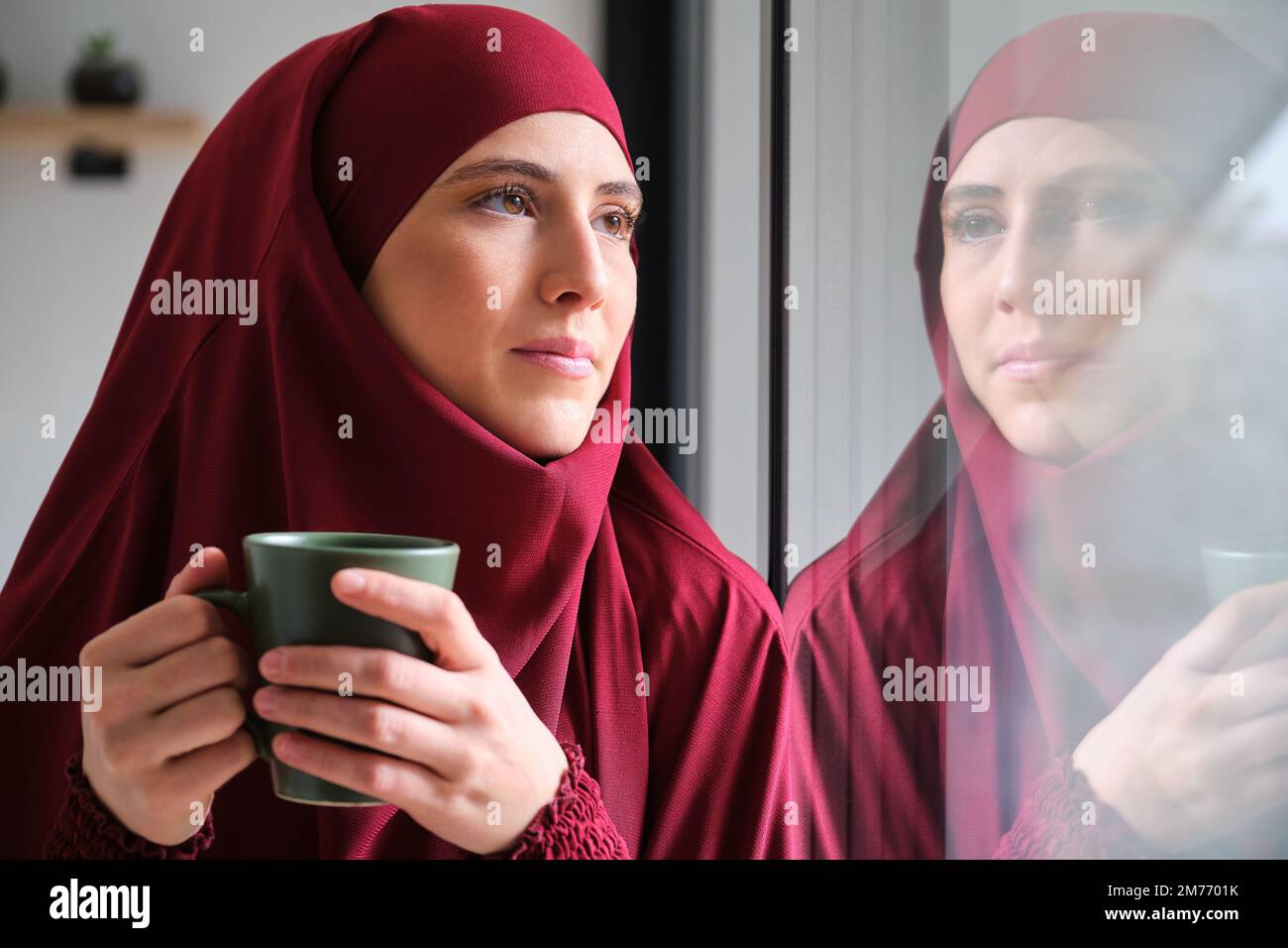 Muslim woman in hijab with a coffee cup looking through a window. Stock Photo