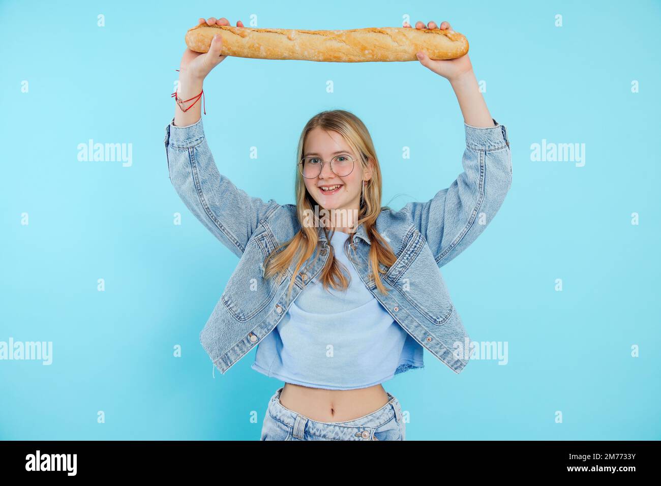 Joyful teenage girl hold over head tasty french baguette on empty blue