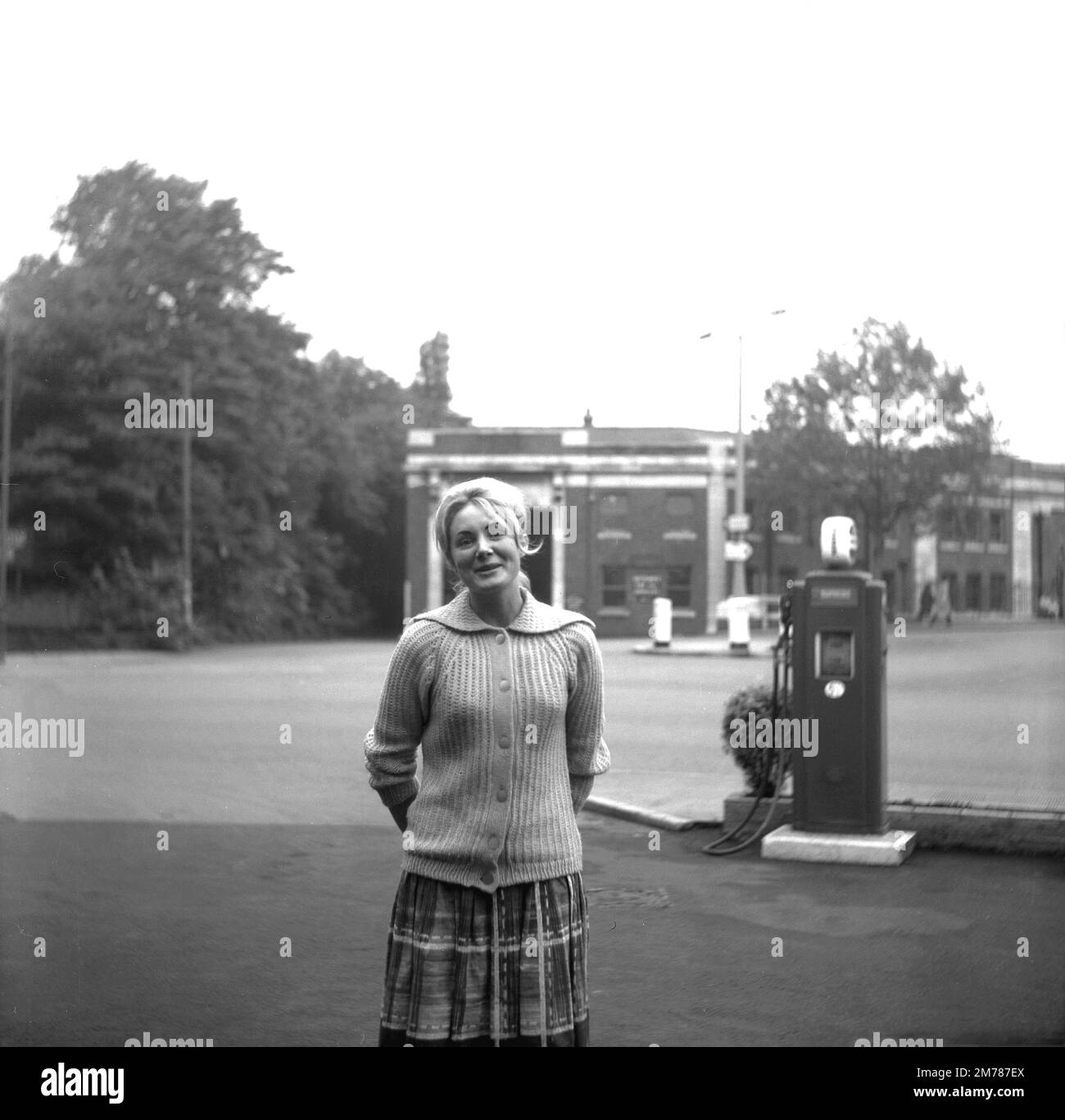 1950s, historical, young woman standing for her photo on the forecourt of the Hargood Motor Co (Parrs wood) Ltd, Didsbury, England, UK. Behind her a petrol pump of the era, possibly a Regent. The bus depot serving Greater Manchester area is seen in the background Stock Photo