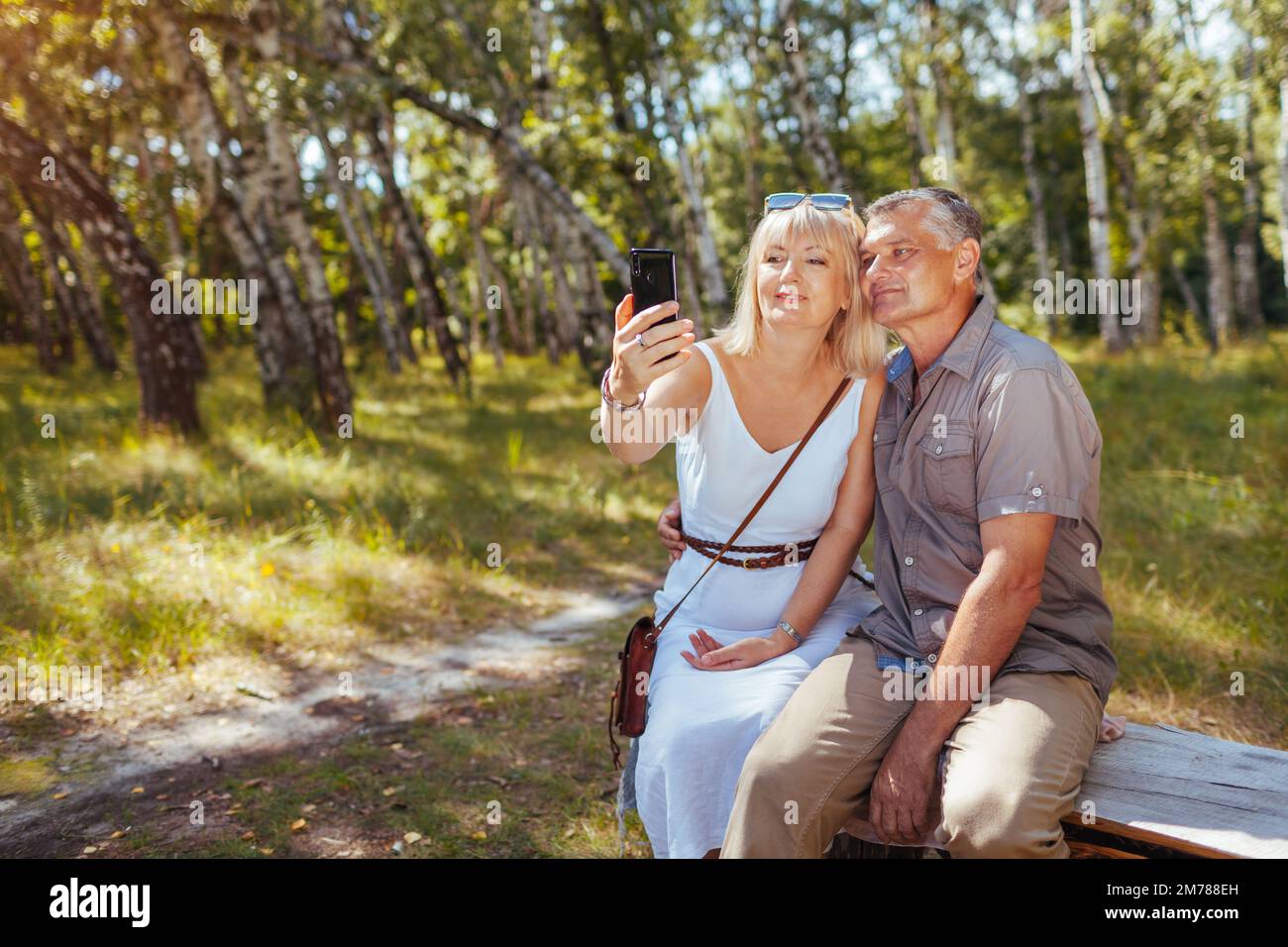 Portrait of senior family couple taking selfie using smartphone in summer forest. Elderly people rest sitting on bench Stock Photo