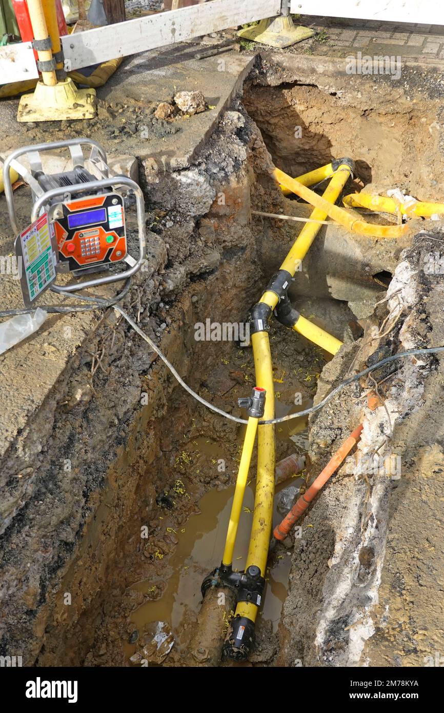 Gas main replacement pit in residential pavement new yellow plastic gas main tubes entering old ageing steel pipes and onwards to each home England UK Stock Photo