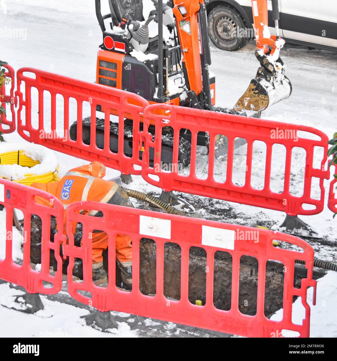 Gas contractor worker bending down snow covered pavement pit trench poor working conditions replacing ageing gas mains in village residential road UK Stock Photo