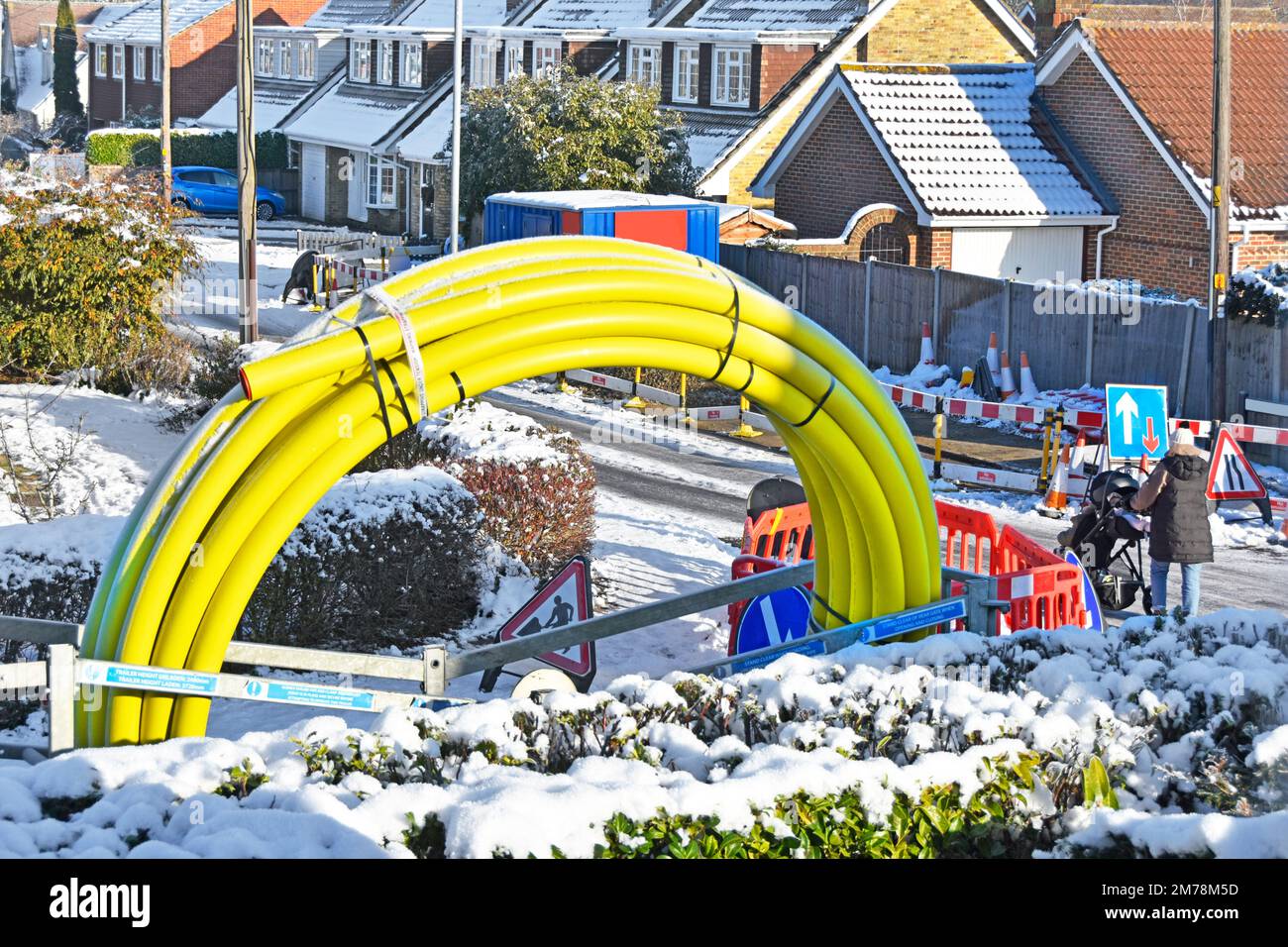 Winter snow mother pushes pram through shut down icy road works in residential village street passing large coiled up yellow gas main pipe England UK Stock Photo