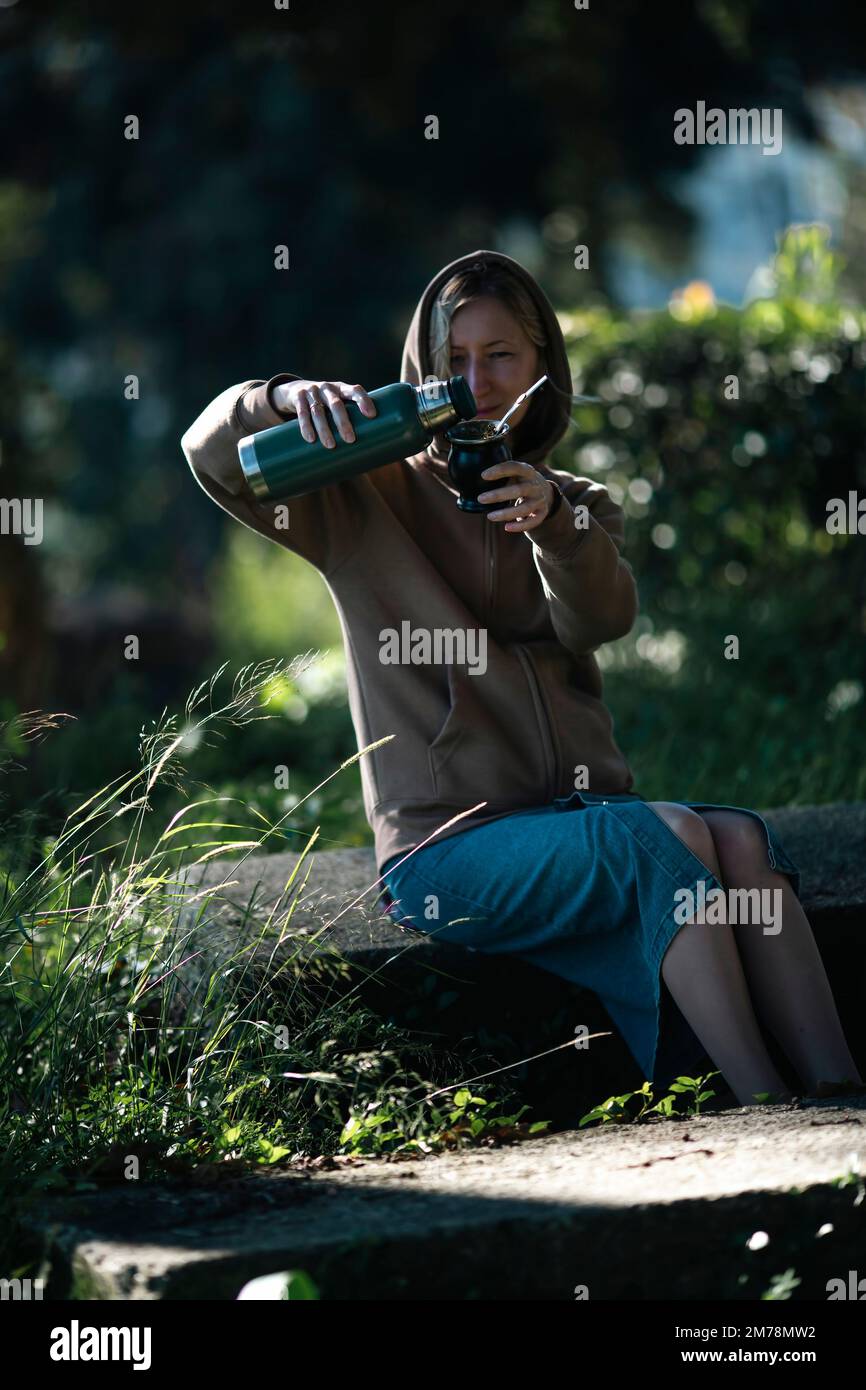 A woman sits in the park pouring hot water from a thermos in a Mate cup. Stock Photo