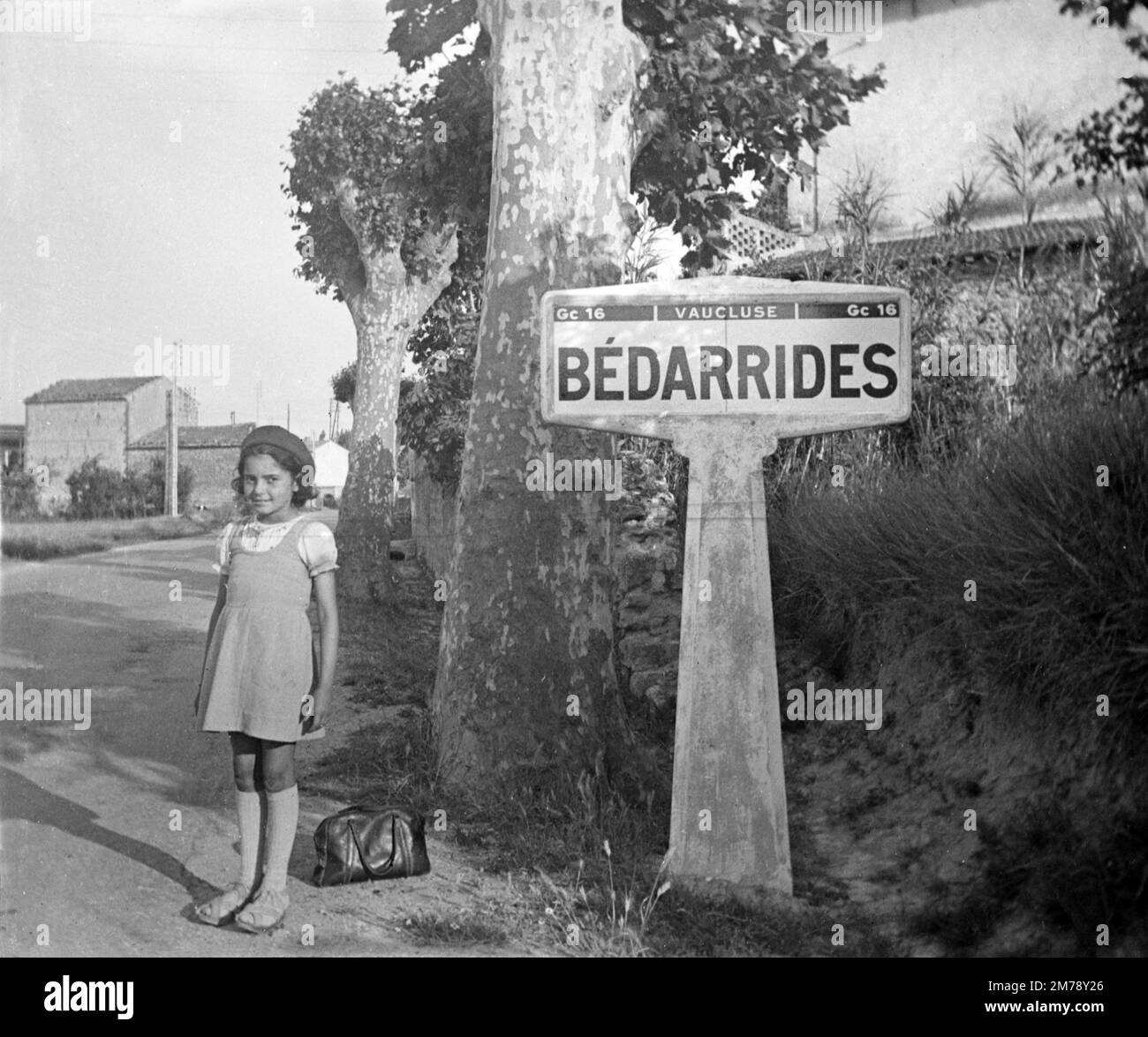 Young French Girl in Beret & 1940s Fashion or Clothes Poses Next to a Village Sign of Bedarrides in the Vaucluse Provence France. Vintage Black and White or Monochrome Photograph 1943 Stock Photo