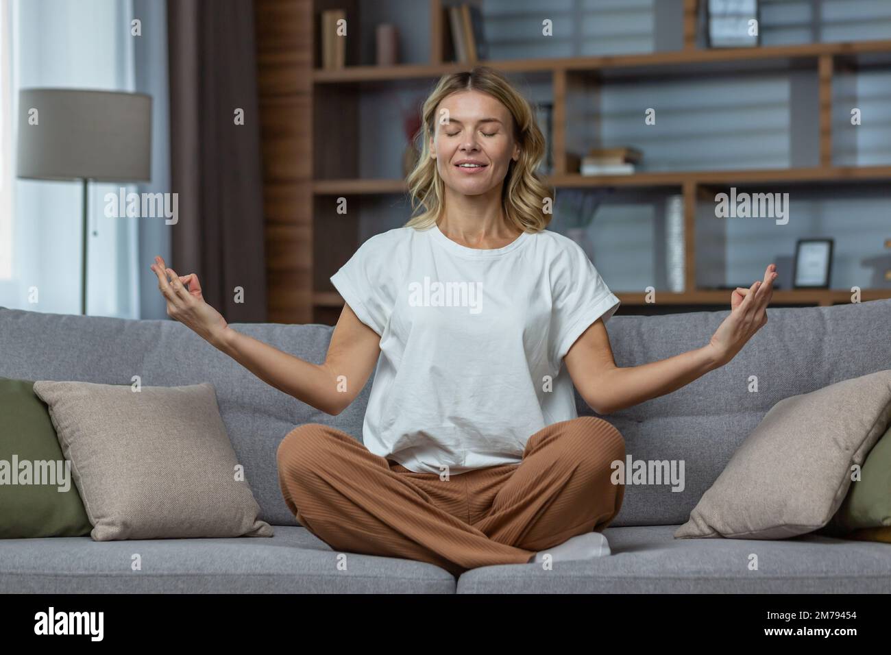 Senior woman at home resting meditating with eyes closed sitting on sofa in living room, housewife mature blonde in lotus pose smiling. Stock Photo