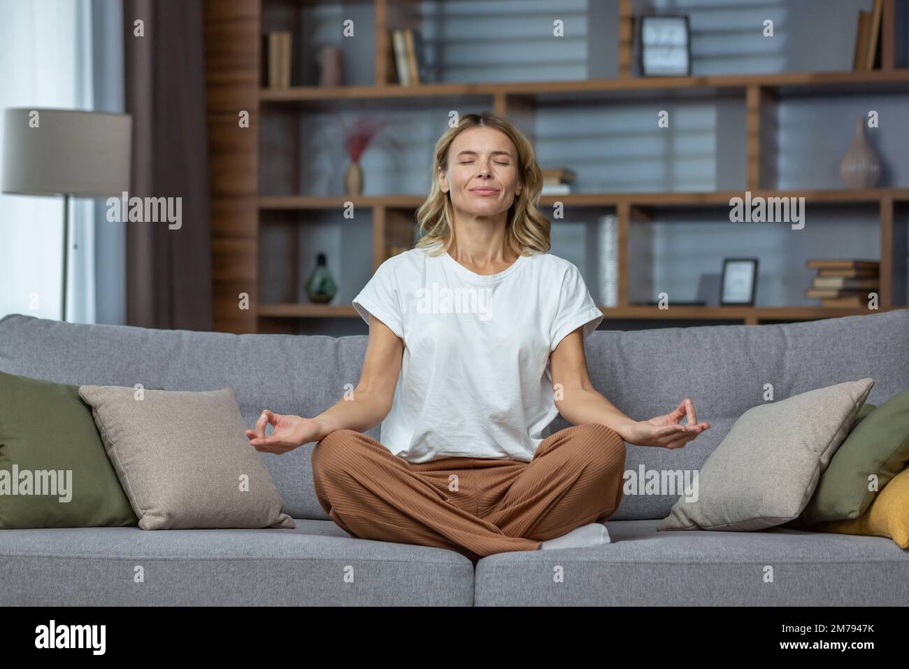 Senior woman at home resting meditating with eyes closed sitting on sofa in living room, housewife mature blonde in lotus pose smiling. Stock Photo