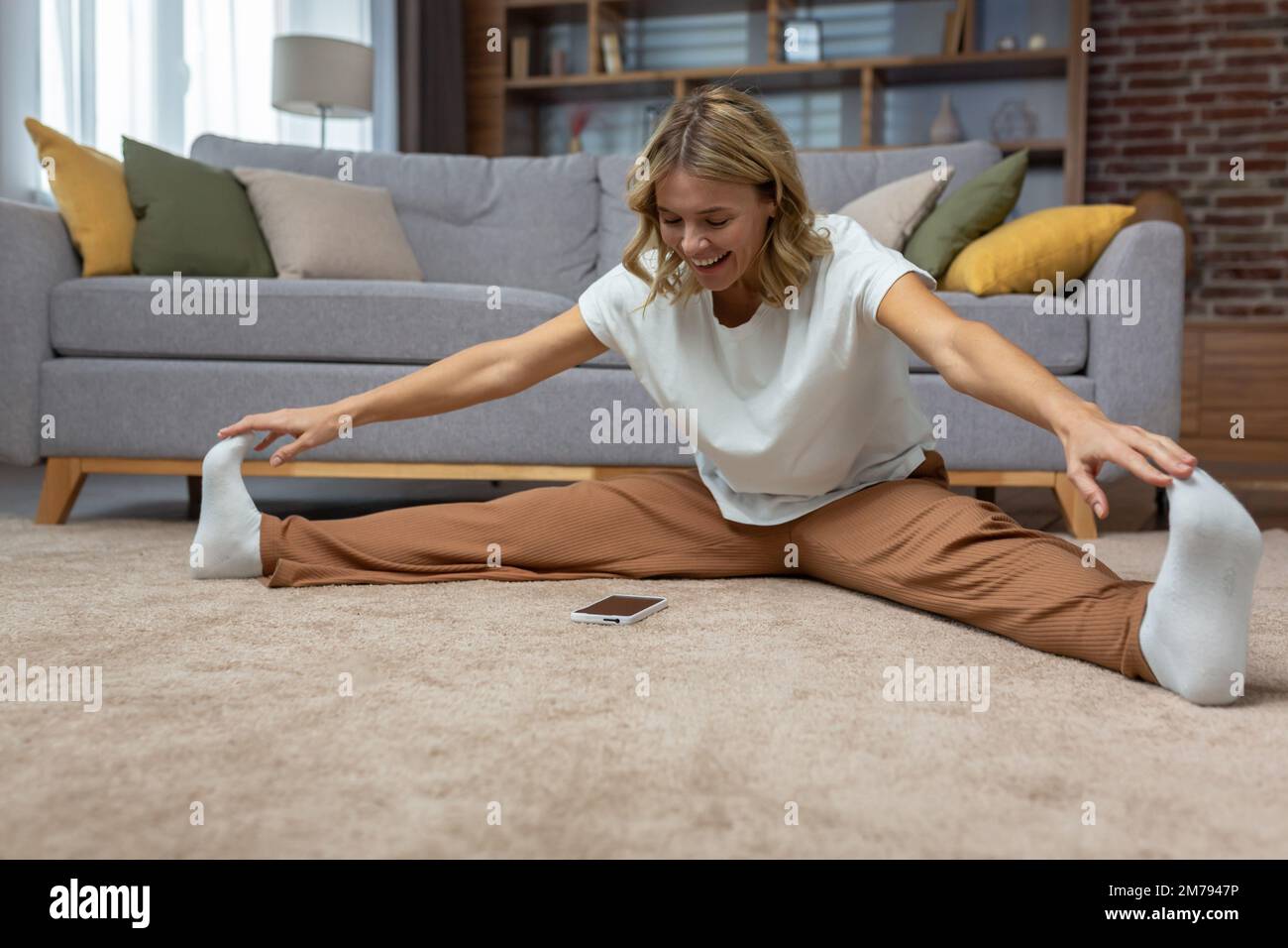Smiling active blonde senior woman on floor doing stretches and exercises, using phone and online workout app at home in living room. Stock Photo
