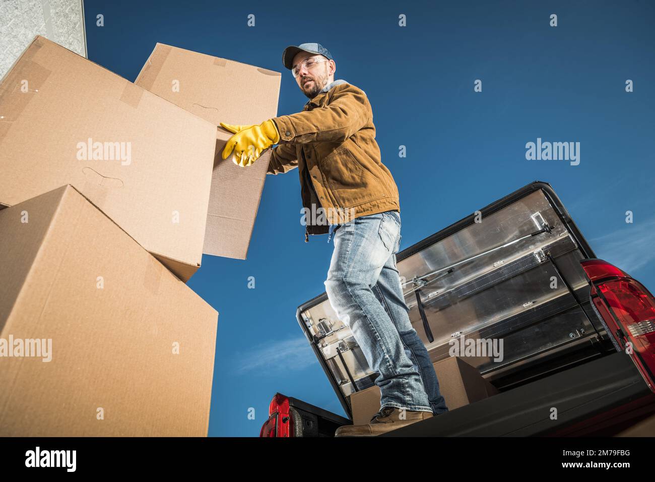 Moving Company Worker Loading Big Cardboard Boxes on the Cargo Bed of His Pickup Truck. Relocation Services Theme. Stock Photo