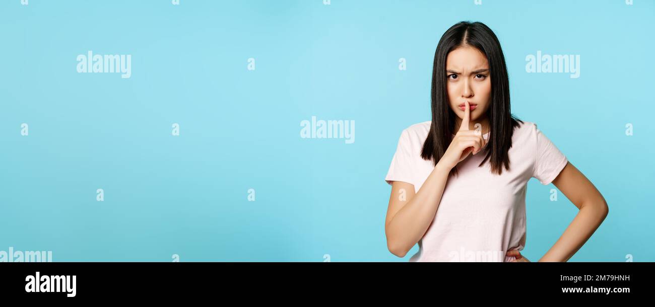 Angry asian woman shushing, frowning in disapproval and hushing, taboo sign, silence gesture, standing over blue background Stock Photo
