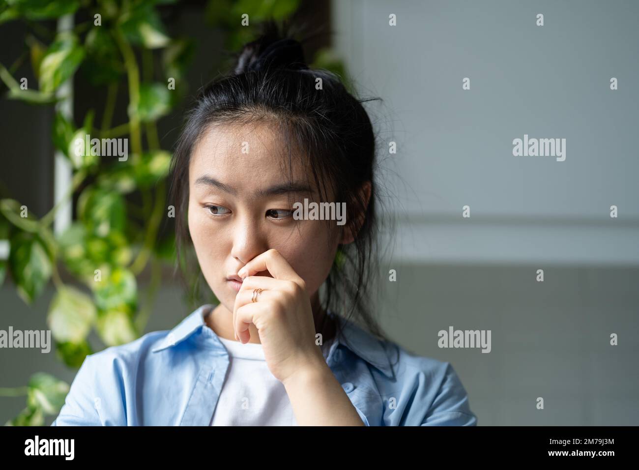 Worried pensive Asian woman touching lips nervous stands in apartment near closet with houseplants Stock Photo