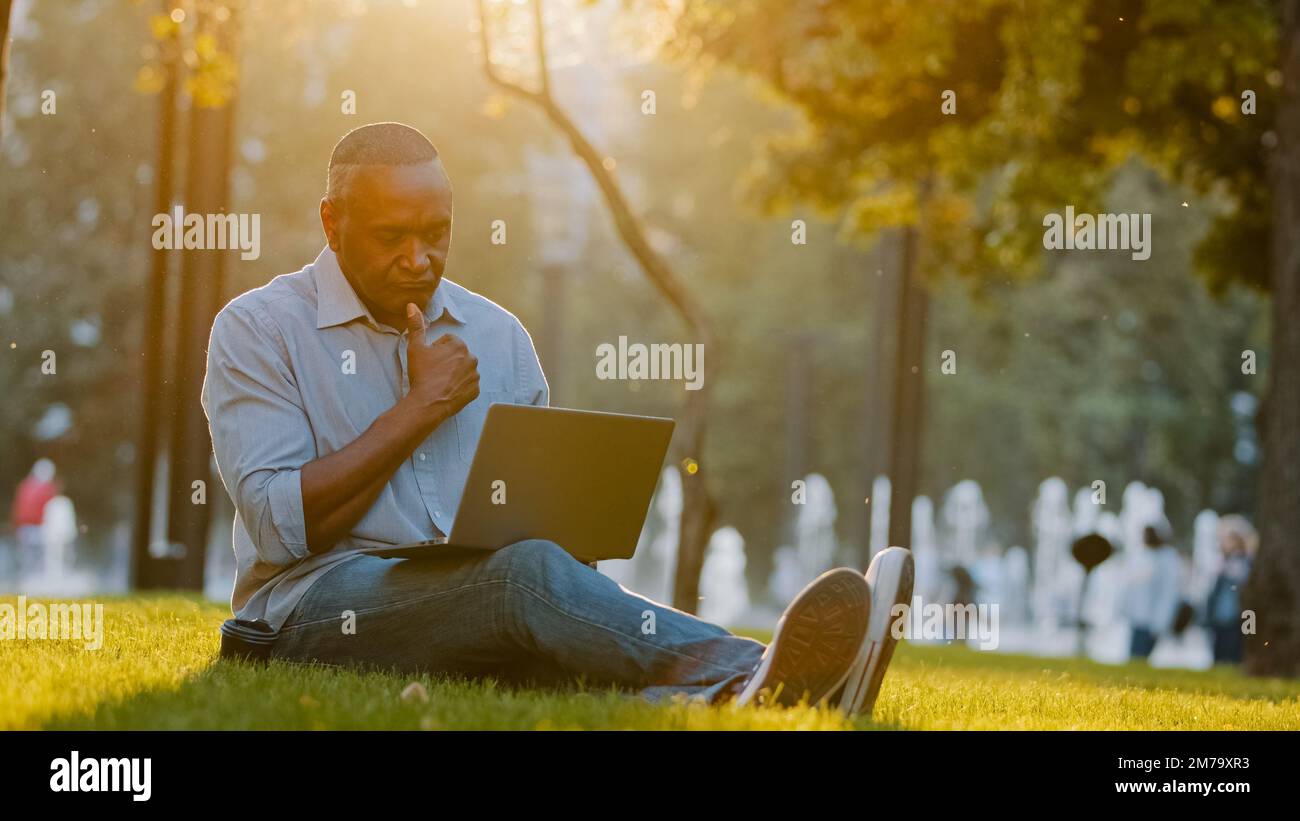 Elderly adult freelancer author writer journalist or blogger african american man sitting outdoors on grass using laptop wireless Internet on modern Stock Photo