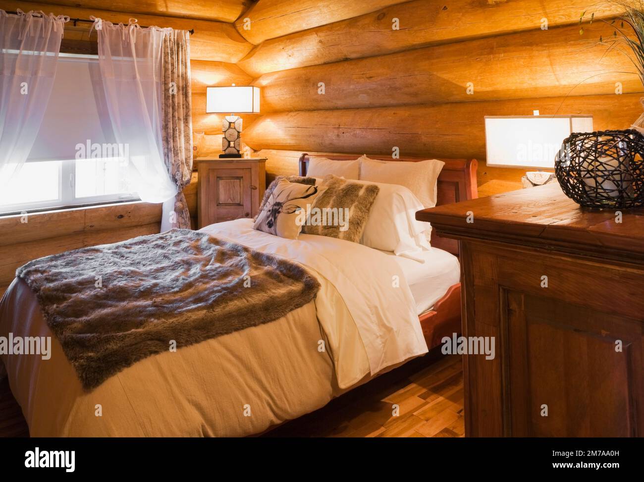 Queen size bed and wooden night tables with illuminated lamps in guest bedroom on ground floor inside luxurious Scandinavian log home. Stock Photo