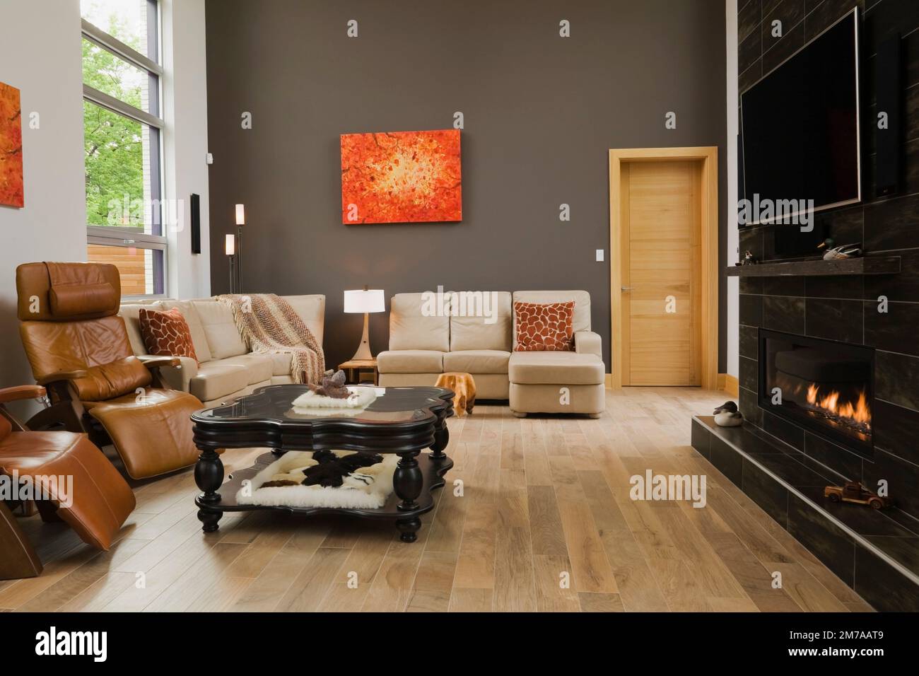 Caramel leather high back reclining armchairs and tan sofas with black wooden glass top coffee table and lit propane gas fireplace in living room. Stock Photo