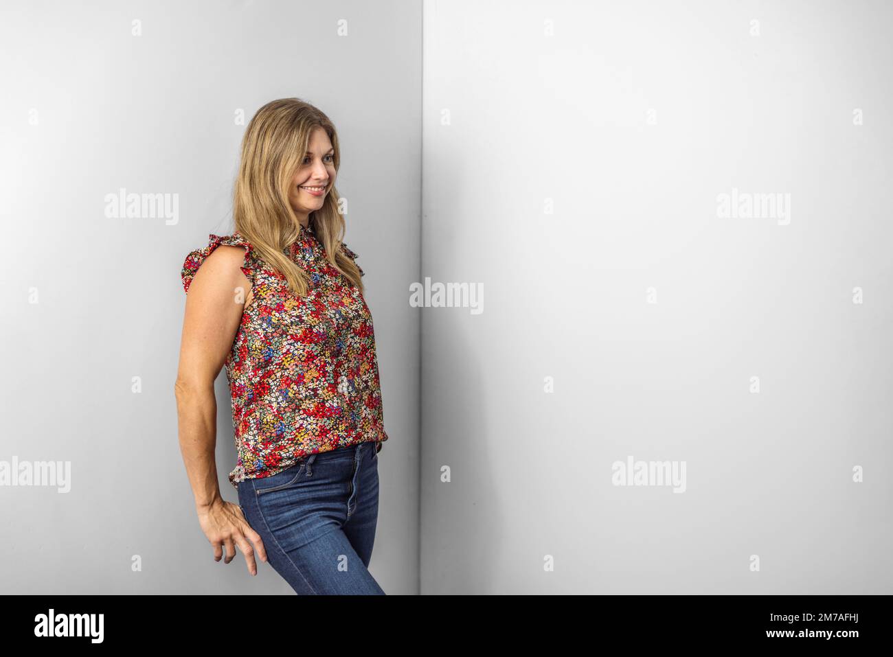 a beautiful caucasian woman in her forties in a casual floral top and denim jeans standing against a white wall with copy space Stock Photo