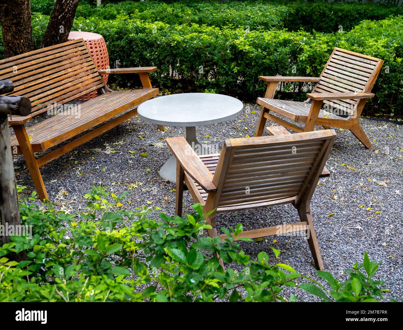 Wood table set in the garden. White empty round table around with empty wooden armchairs seat and wood bench chair on the gravel ground near the green Stock Photo