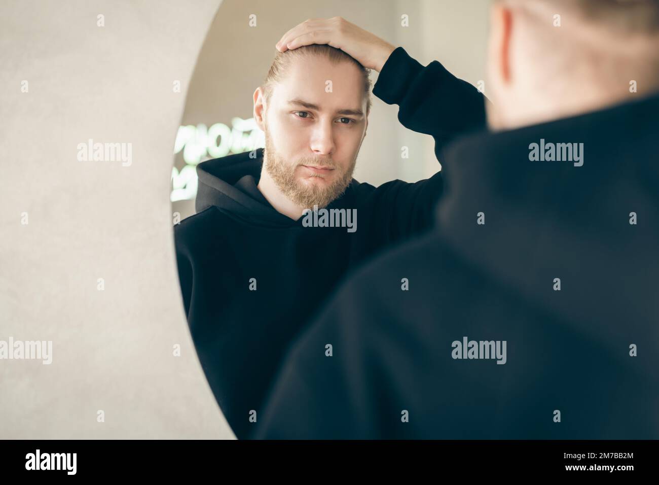 An attractive young man with a beard looks at himself in the mirror. Stock Photo