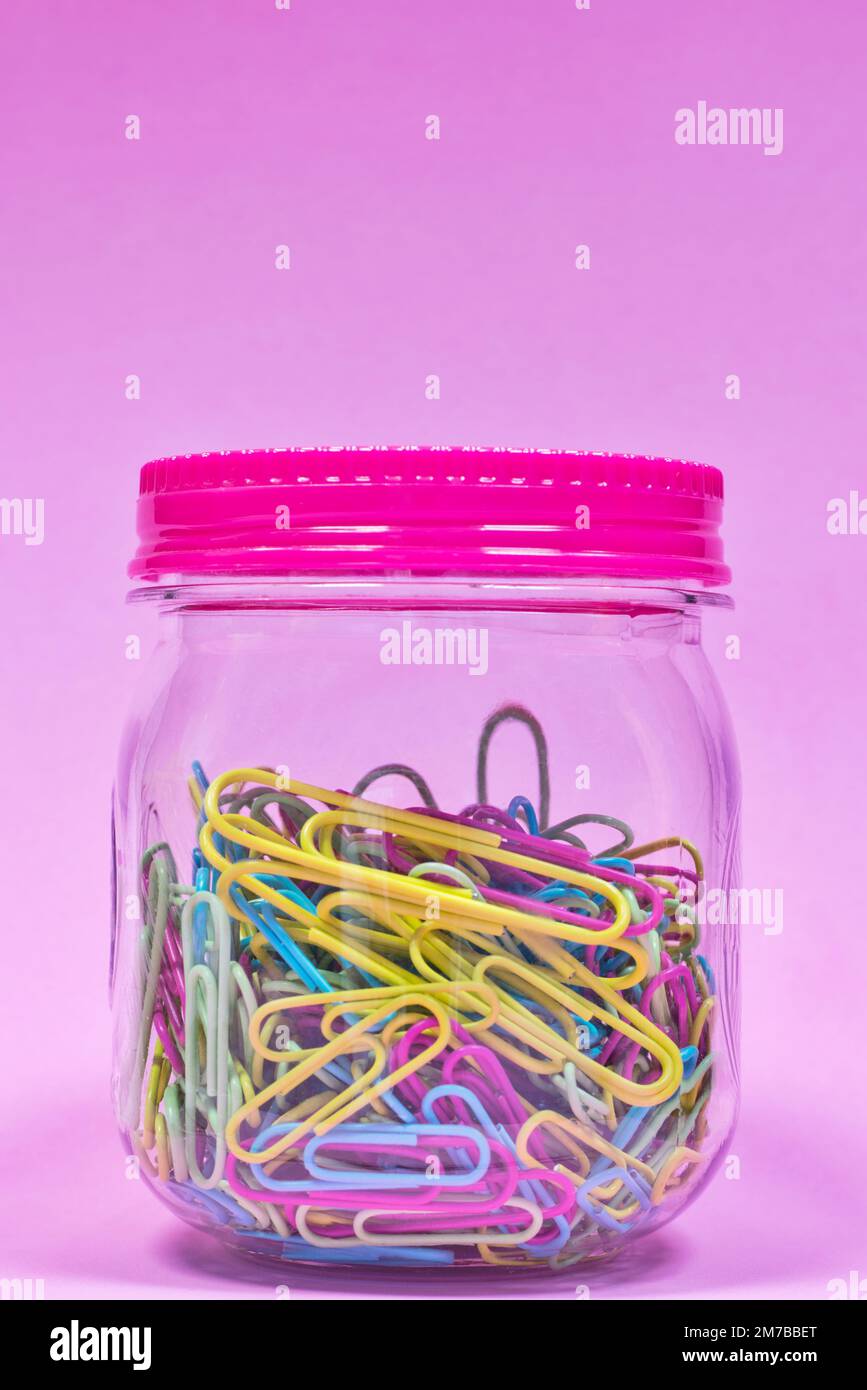 Colorful paperclips in a plain glass jar and sealed lid with pink background and copy space above, creative concept. Stock Photo