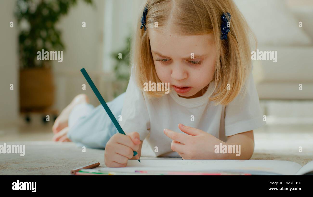 Cute concentrated Caucasian little kid girl draw color pencil lying on floor at home play alone. Talented preschool child daughter kid baby coloring Stock Photo