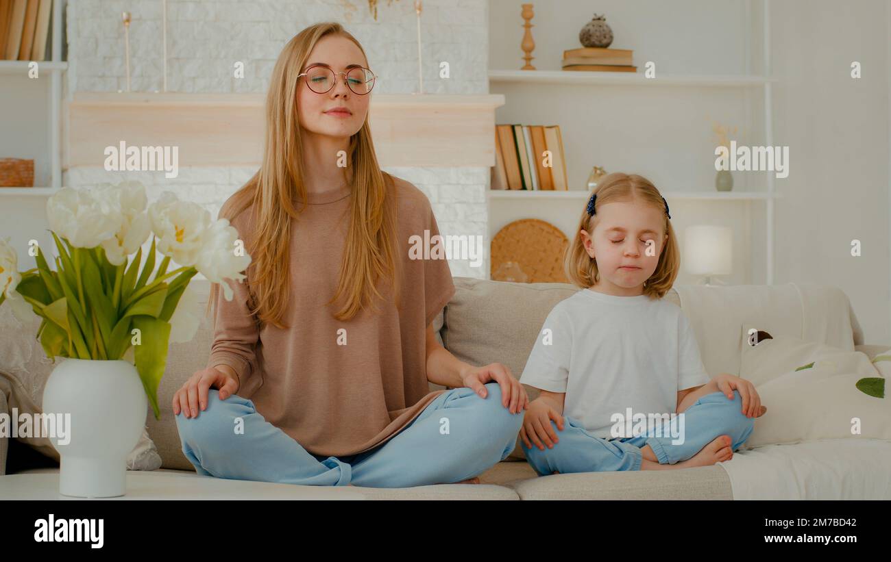Caucasian woman mom with little child girl sitting home at couch in lotus position closed eyes meditate together. Family meditating mother teach Stock Photo