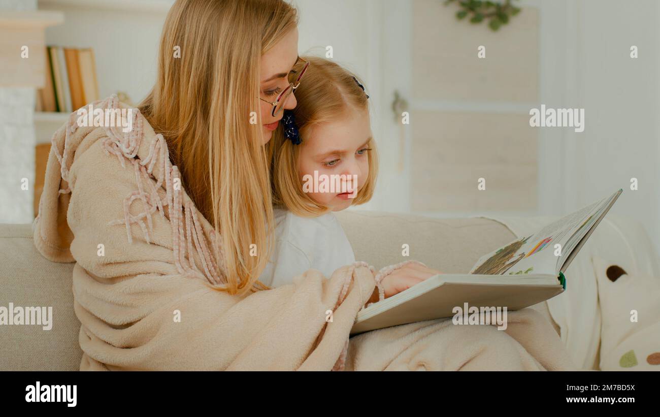 Little small Caucasian daughter child girl kid baby with mother in living room reading literature looking at photo album mom read book showing Stock Photo