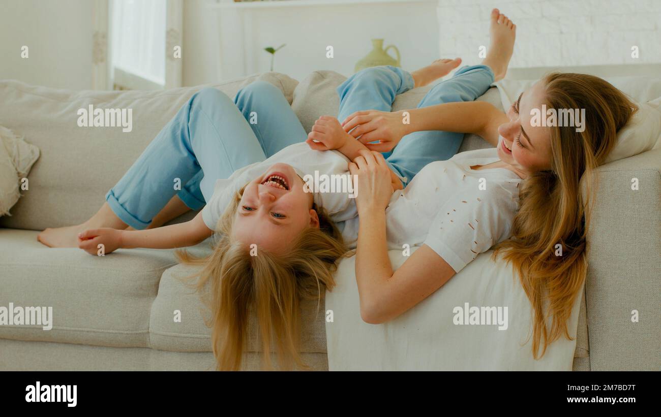 Caucasian mother mom with daughter lying on couch sofa in living room mommy tickle kid girl laughing tickling child baby sincere funny laugh enjoy Stock Photo