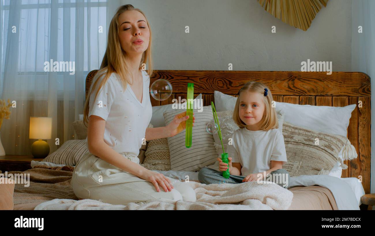 Caucasian mother woman mom with small little child girl kid baby adopted healthy daughter blowing soap bubbles in air in bedroom sitting on bed Stock Photo