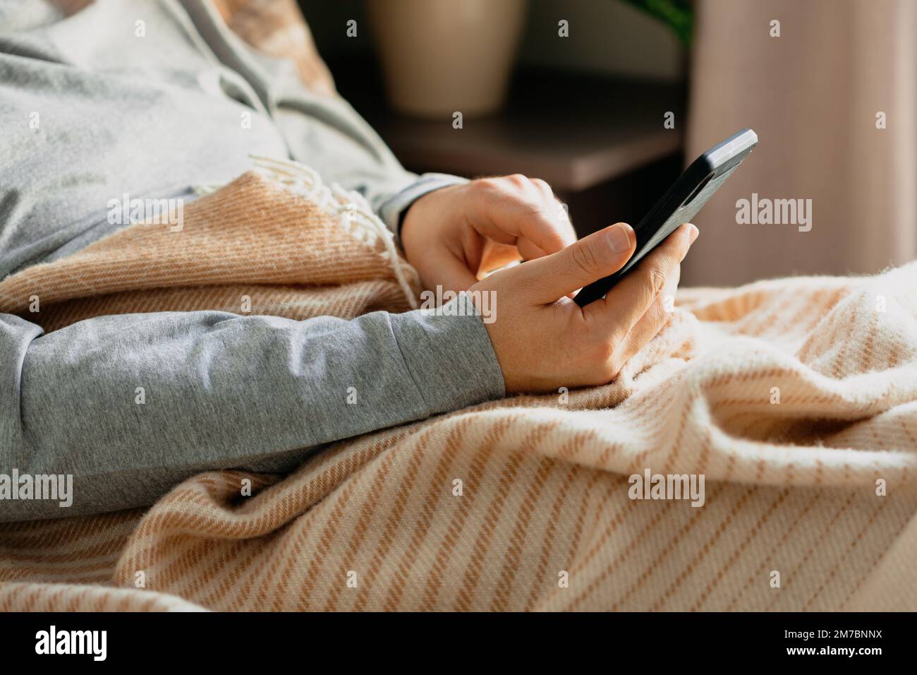 a caucasian man relaxing, using smart phone lying in bed under throw at home Stock Photo