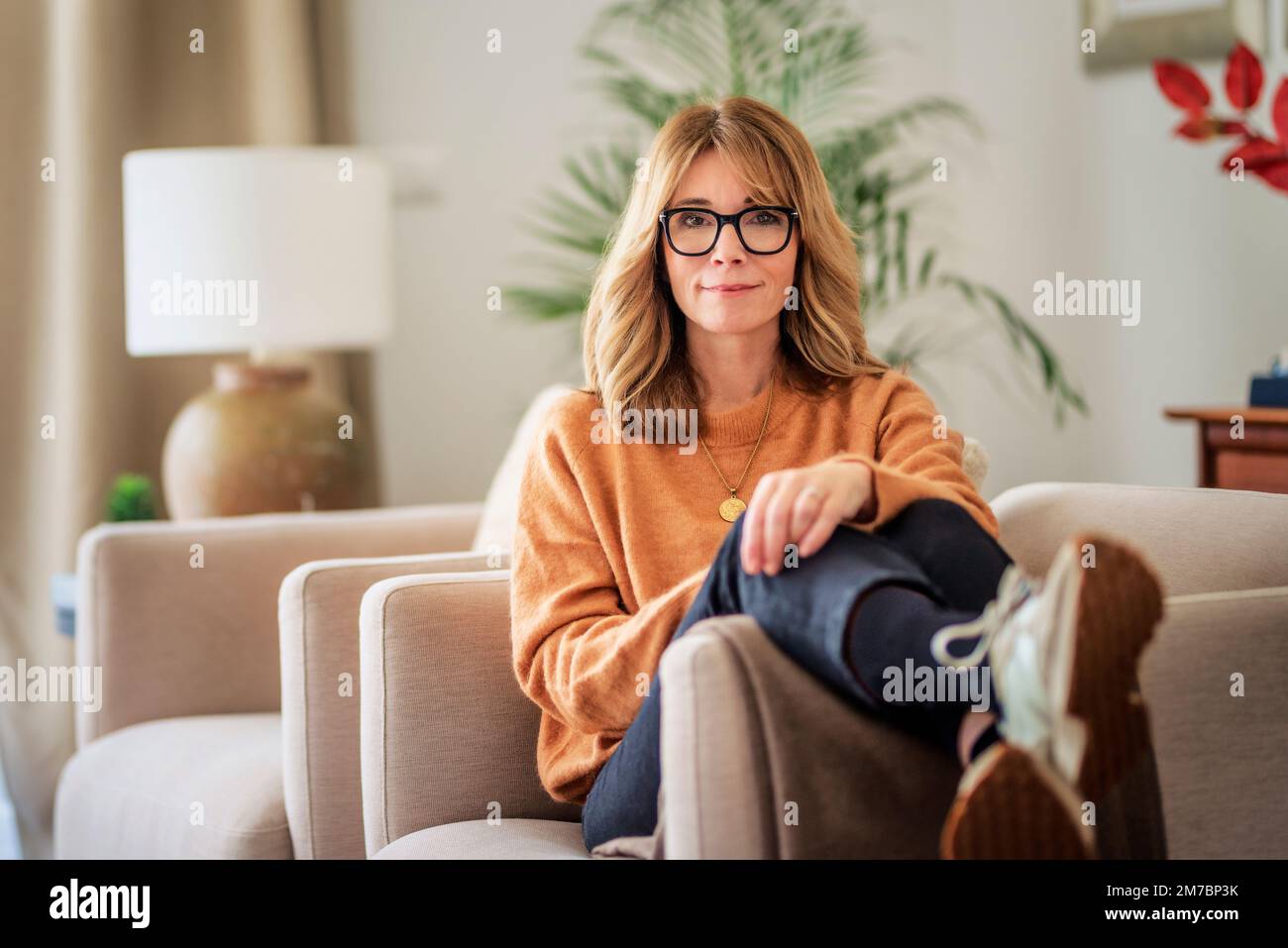 Portrait of attractive middle aged woman relaxing in an armchair at home. Blond haired female wearing eyeglasses and sweater. Stock Photo