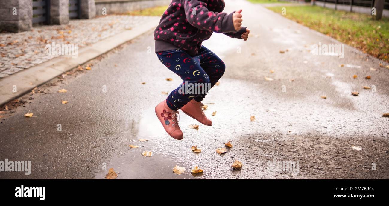 A child jumps over a puddle of water and leaves - the road in the background Stock Photo