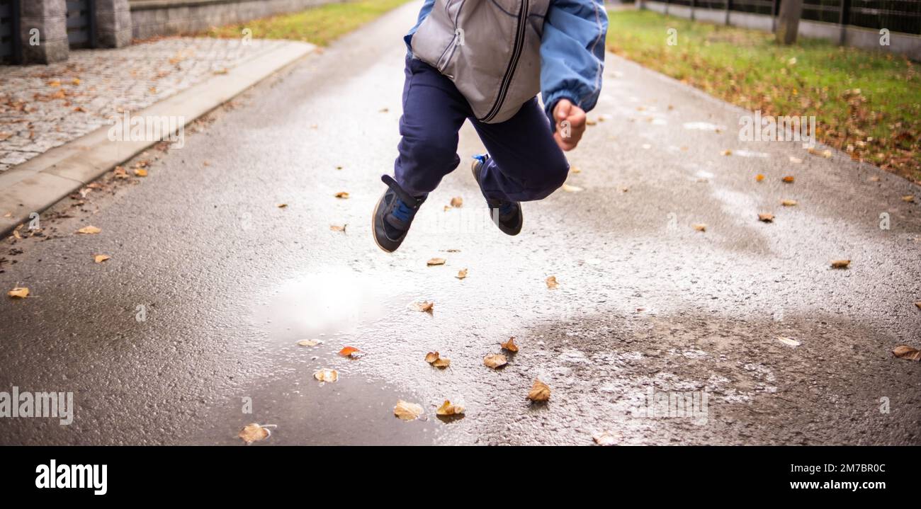 A child jumps over a puddle of water and leaves - the road in the background Stock Photo