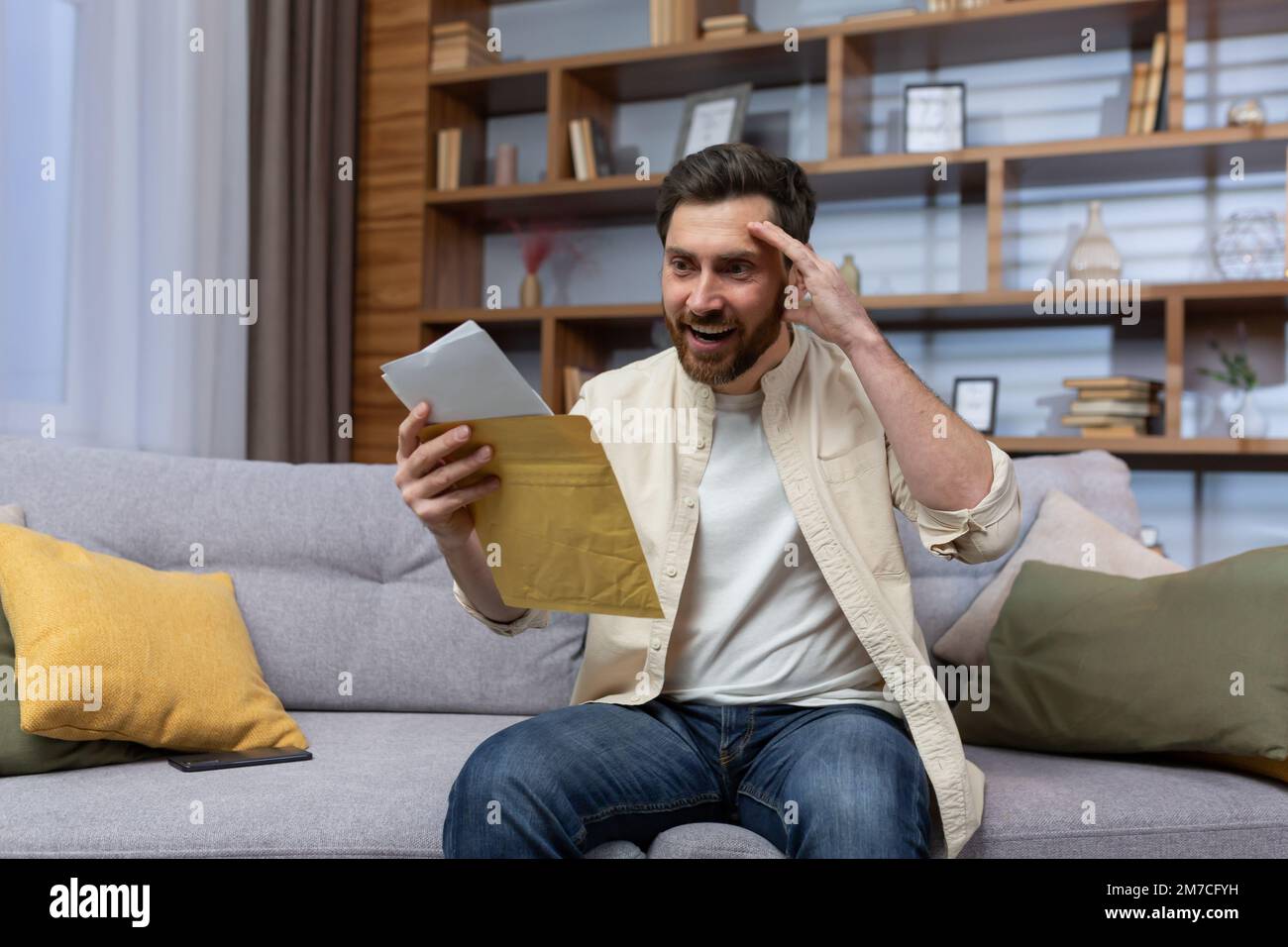Joyful man at home holding envelope with notification message reading and smiling happy with news sitting on sofa inside living room. Stock Photo
