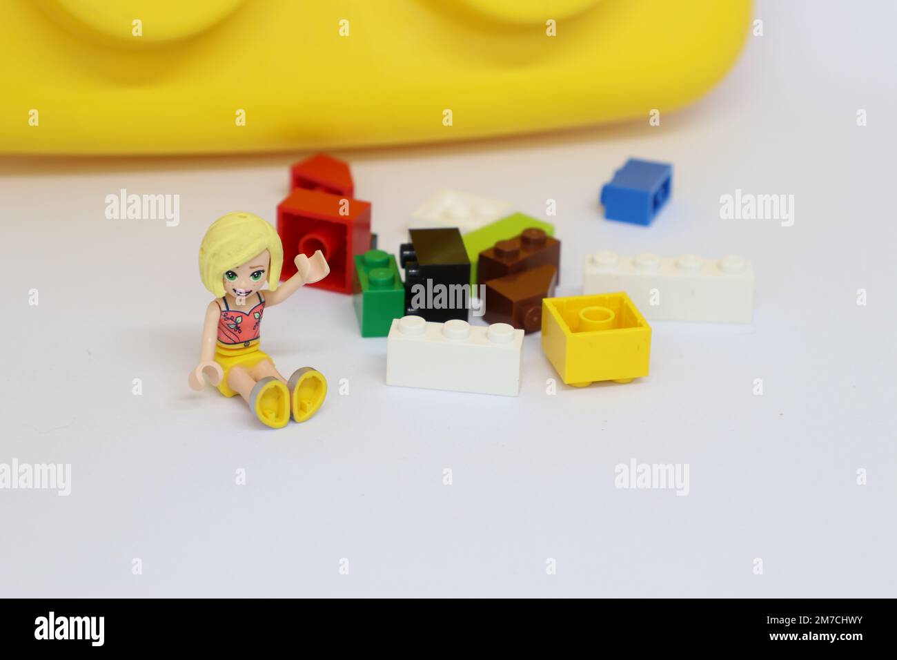 A girl with blonde hair is waving. She is sitting next to scattered lego blocks ann a giant lego block in the background. Stock Photo