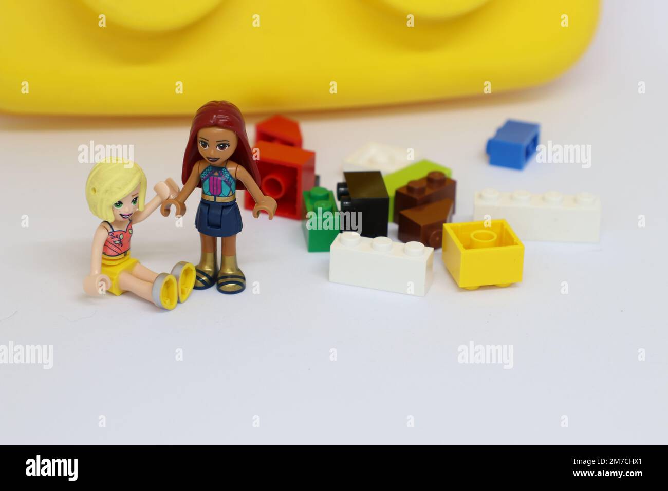 Lego figure girls holding hands next to scattered lego blocks with a giant lego block in the background. Stock Photo