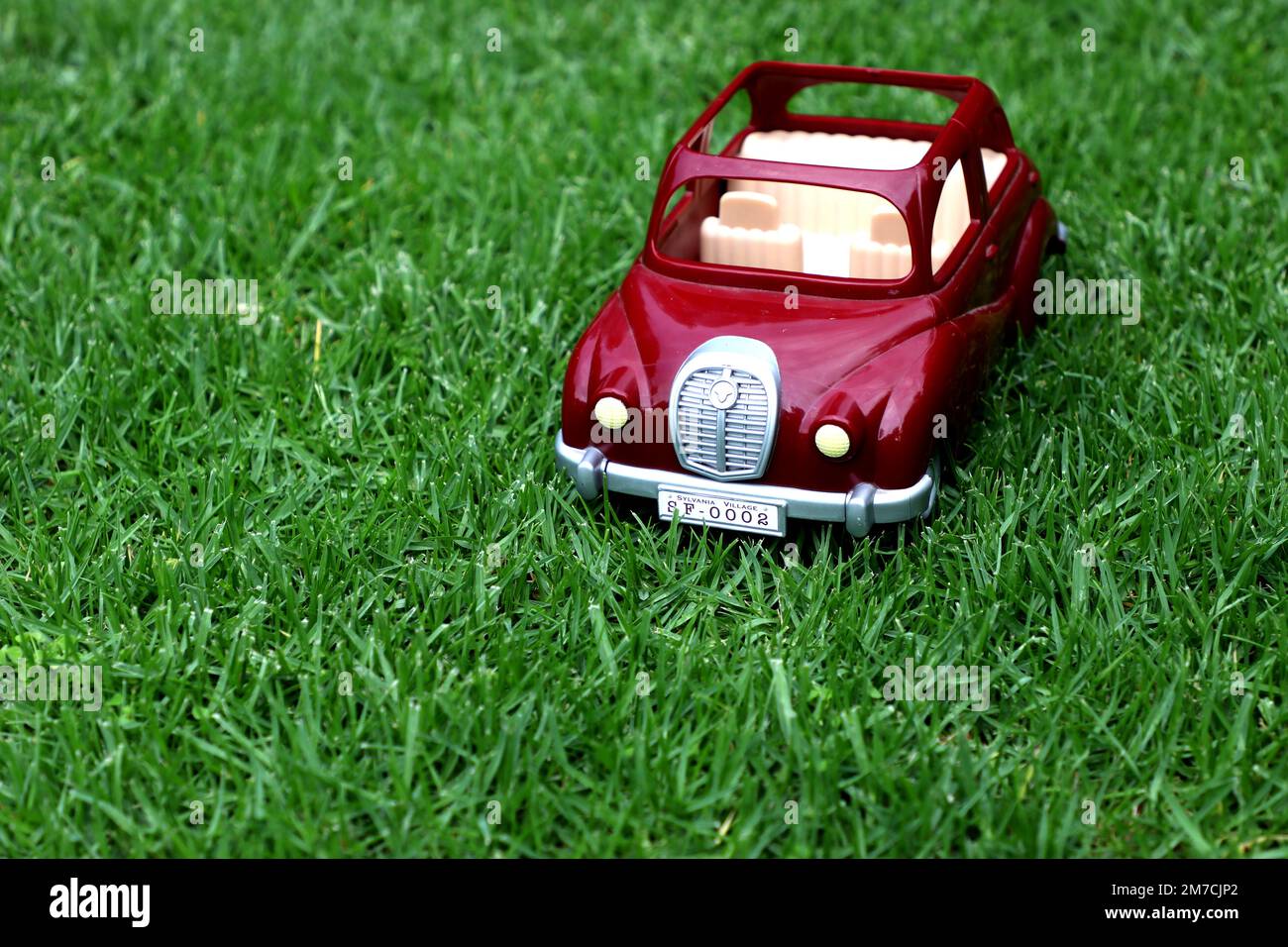 A classic burgundy plastic toy car with the roof down, a silver bumper and fender and white seats sitting on grass Stock Photo