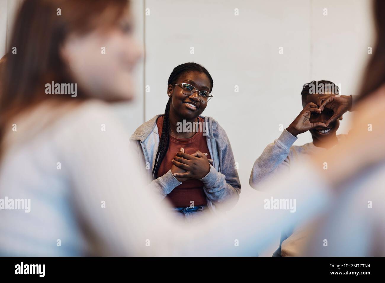 Smiling teenage girl sitting with hands on chest by male friend gesturing heart shape and looking at students in group t Stock Photo