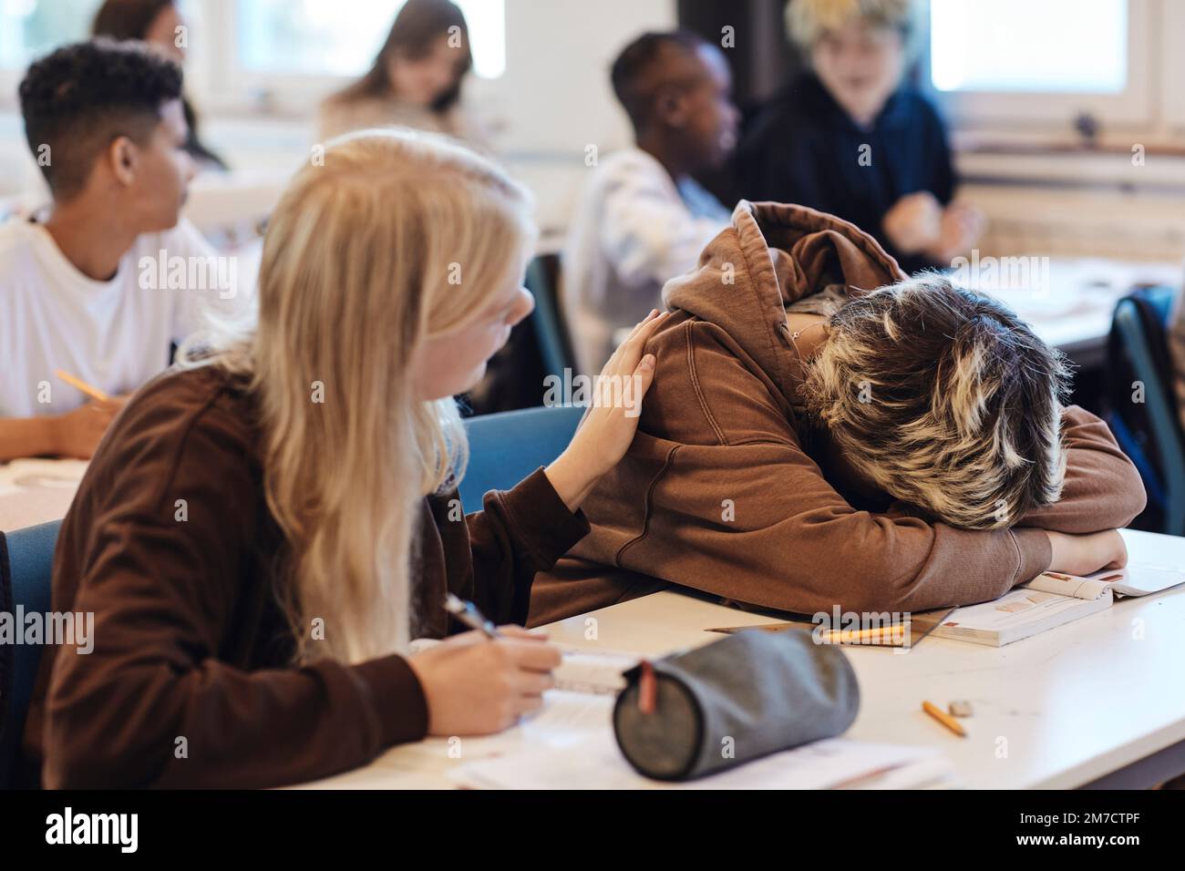 Blond teenage girl consoling depressed female friend resting head on desk in classroom Stock Photo