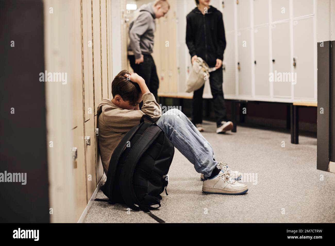Full length of depressed boy sitting with backpack in school corridor Stock Photo