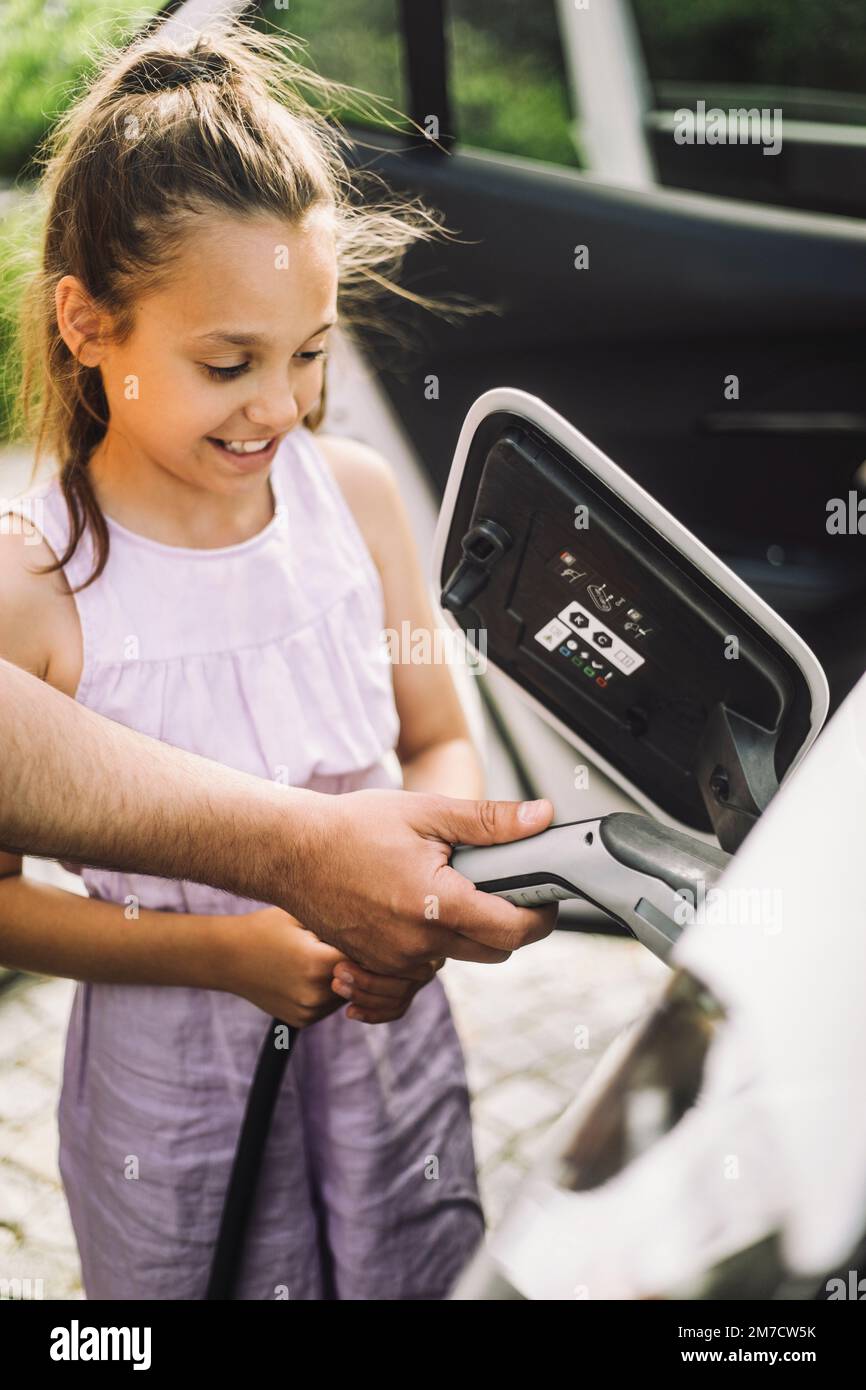 Hand of father teaching daughter to plug cable in electric car Stock