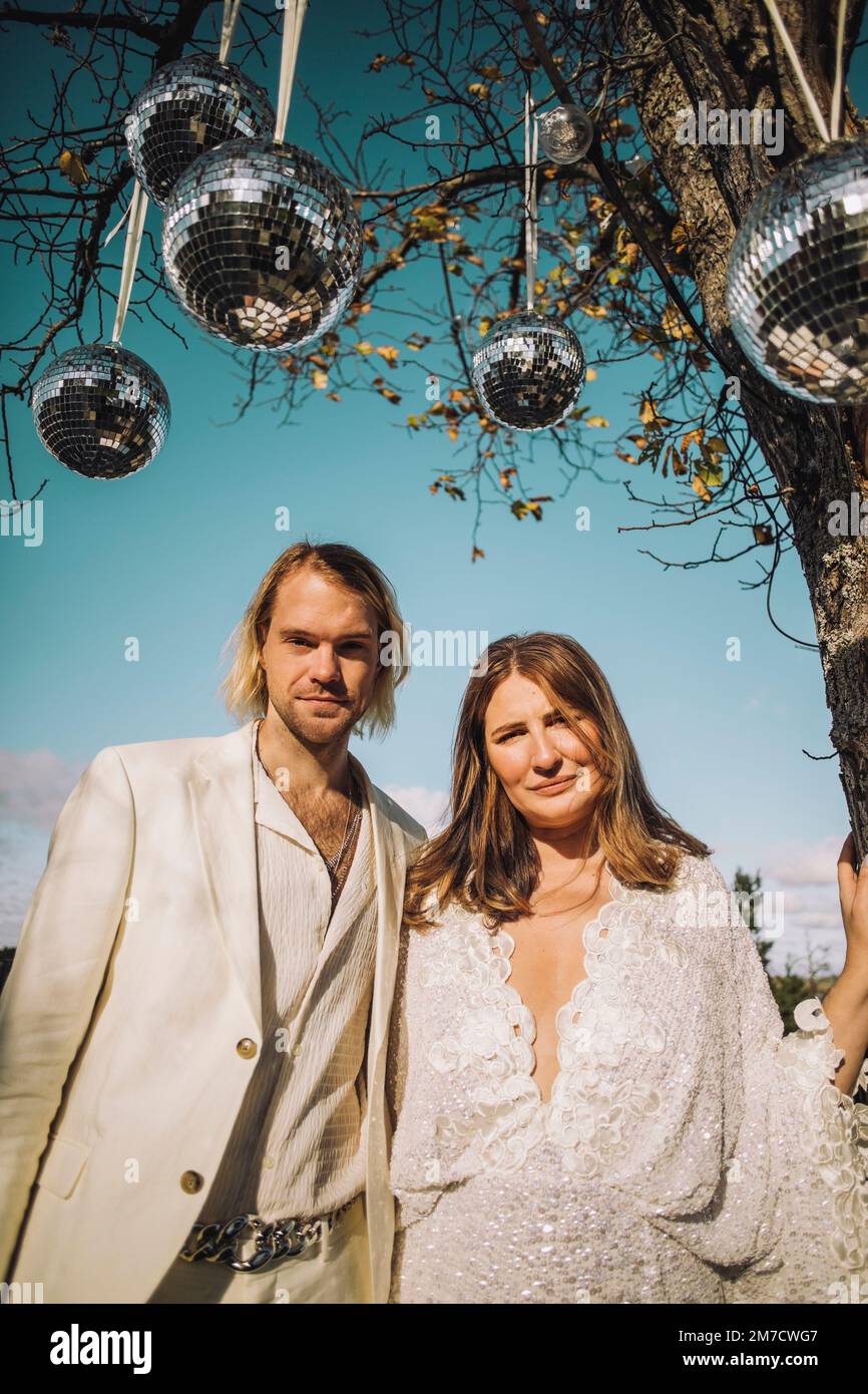 Portrait of newlywed mid adult groom and bride standing under disco balls on sunny day Stock Photo