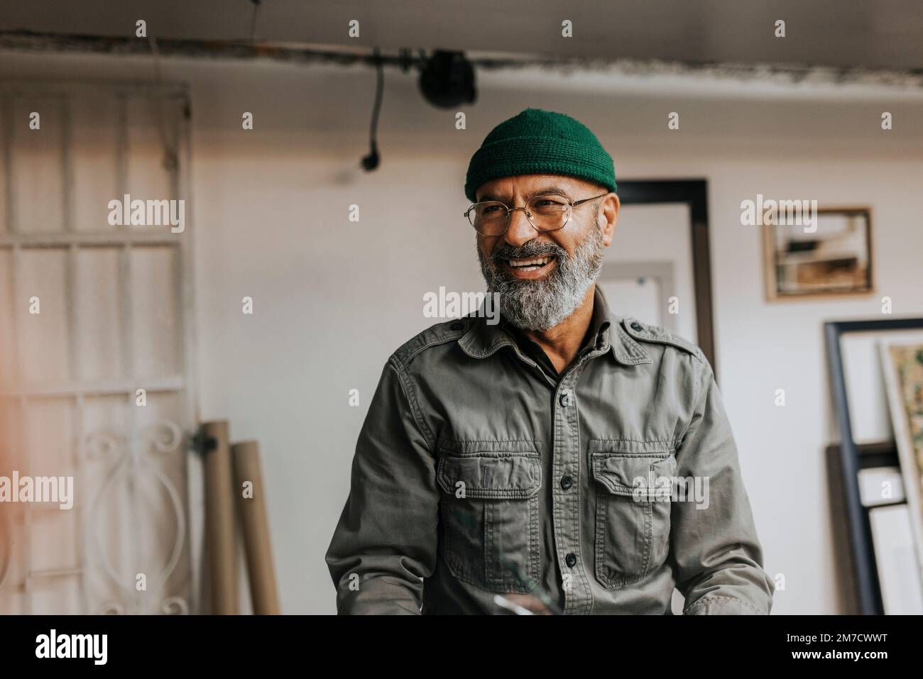 Happy bearded craftsman wearing eyeglasses and knit hat in workshop Stock Photo